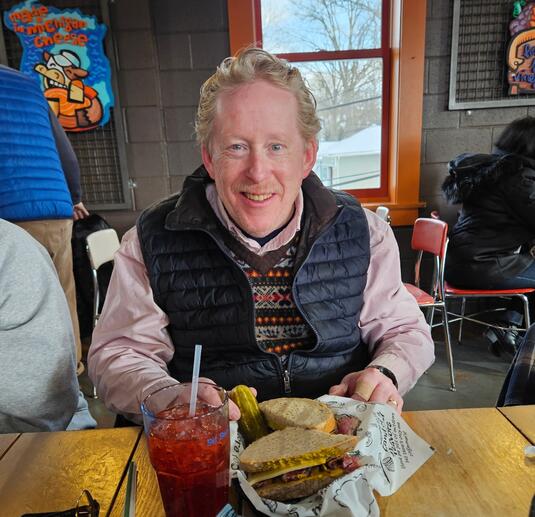 A man smiles at a restaurant with a sandwich and icy drink.