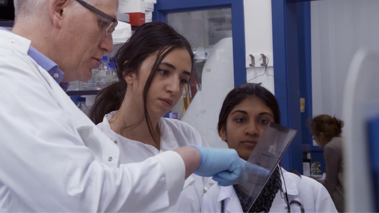 Three researchers in white lab coats examine a clear plastic sheet together in a laboratory.