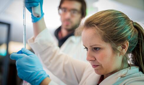Woman wearing gloves and a white coat holding a long pipette with a man in a lab coat watching in the background.