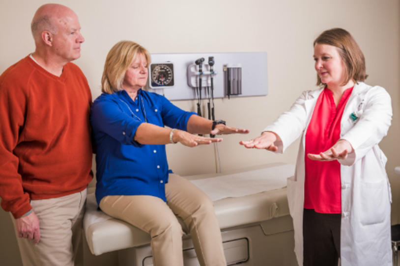 Male care partner alongside female patient undergoing Parkinson's test with her female physician.