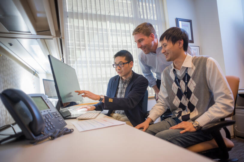 Three male researchers sitting or standing in front of a computer looking at a shared screen. 