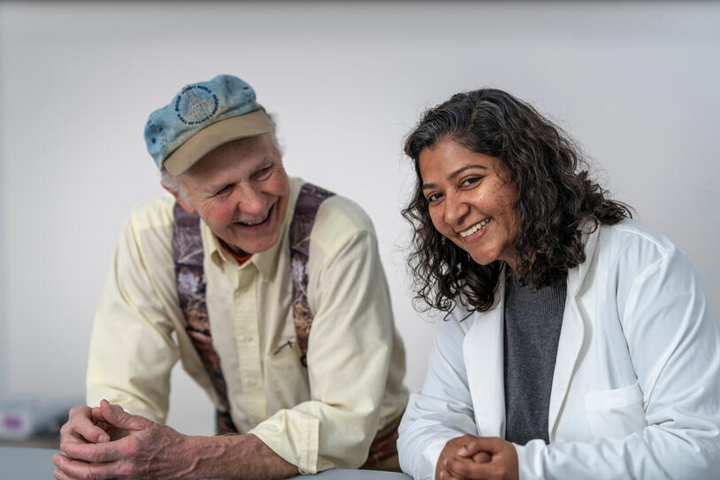 An older patient with a hat and yellow shirt laughs next to his smiling healthcare provider.