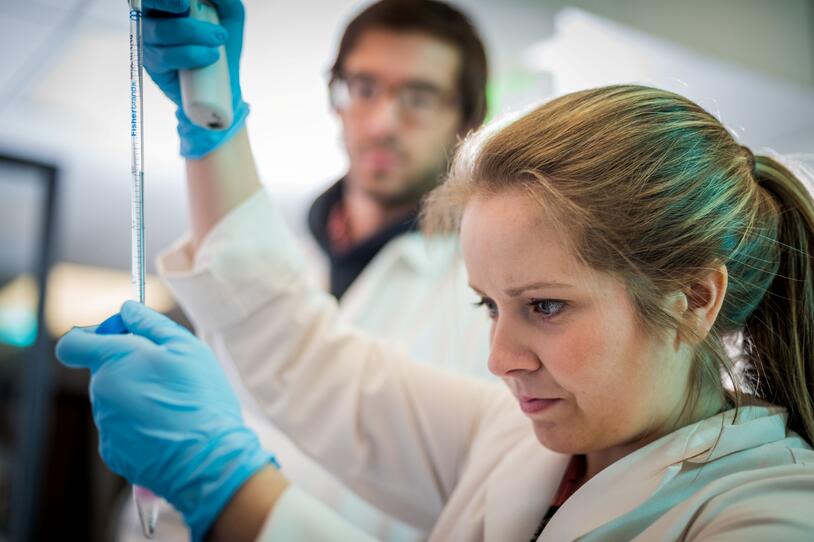 Woman wearing gloves and a white coat holding a long pipette with a man in a lab coat watching in the background.