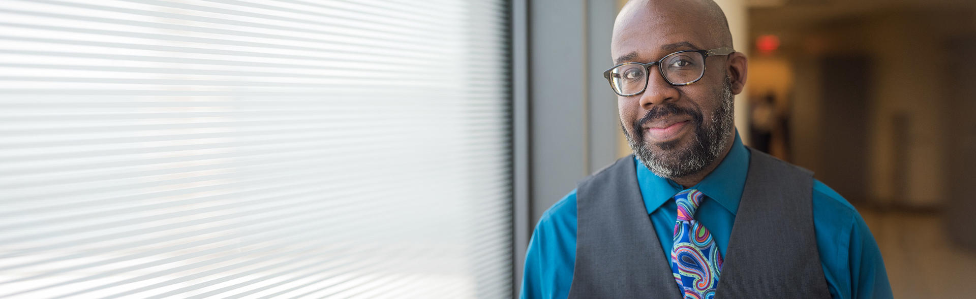 African American male wearing glasses and a blue button down, vest and tie.