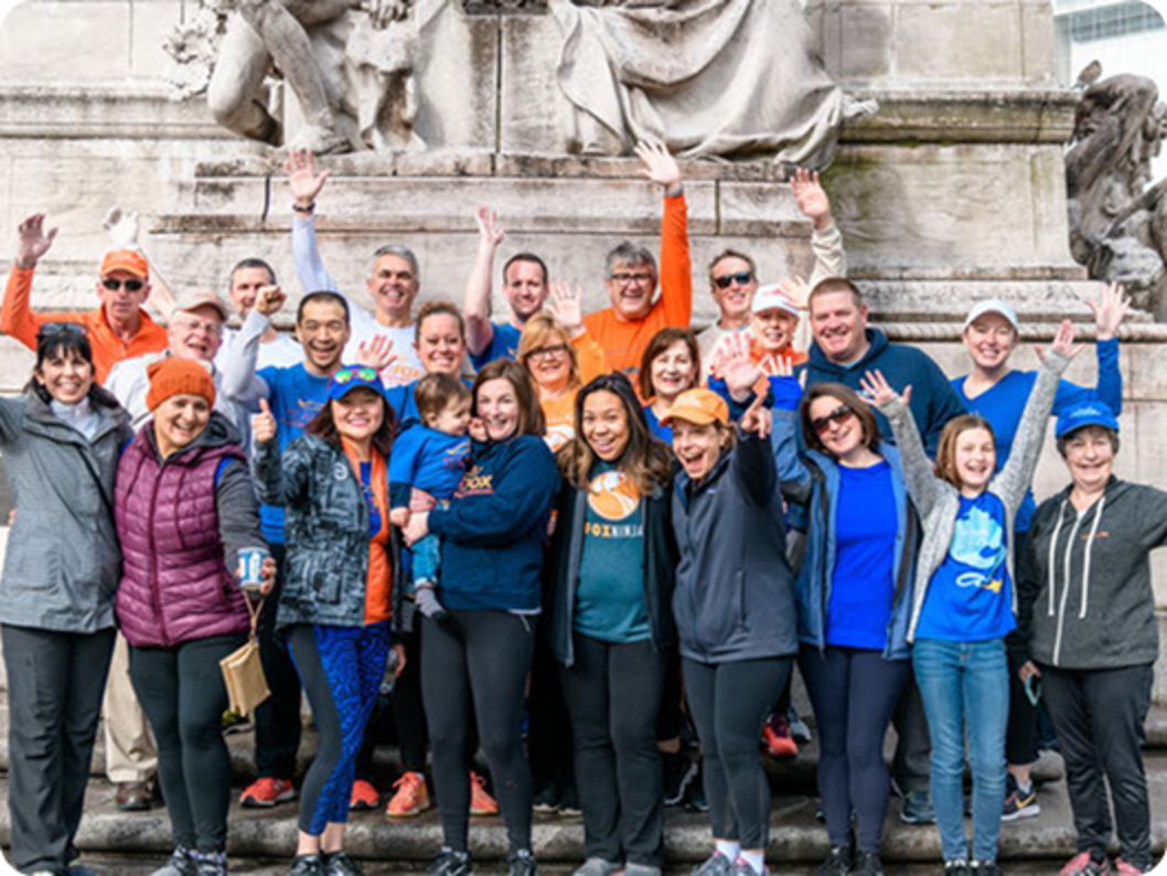 Group of about 20 adults and children in colorful shirts and jackets smiling and waving on stone steps in front of a large monument.