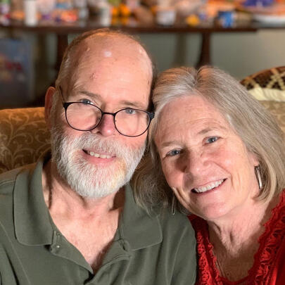Joanie Prentice and her husband sit and smile together on a couch. 