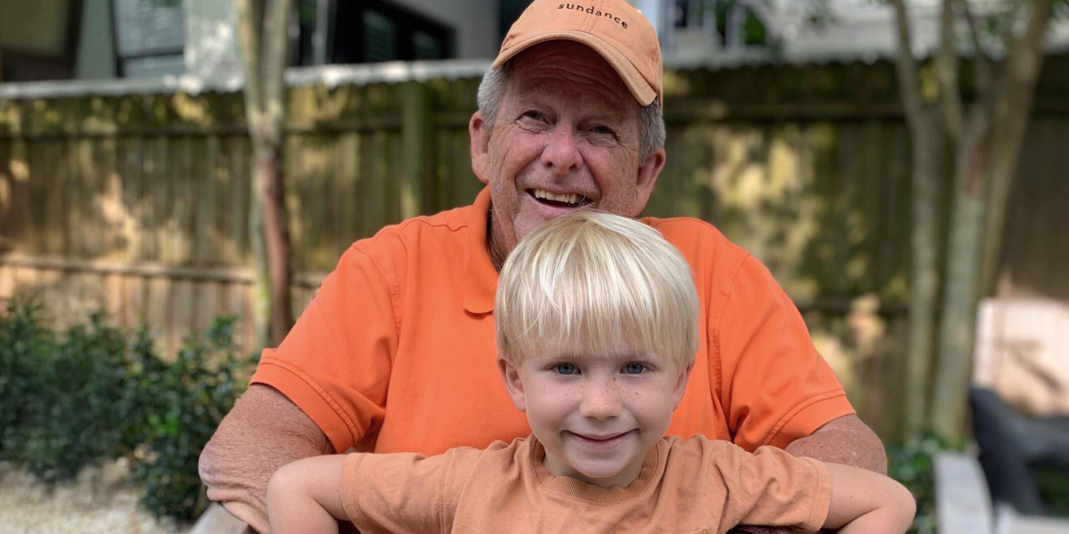A young boy sits on his grandfather’s lap, both wearing orange shirts