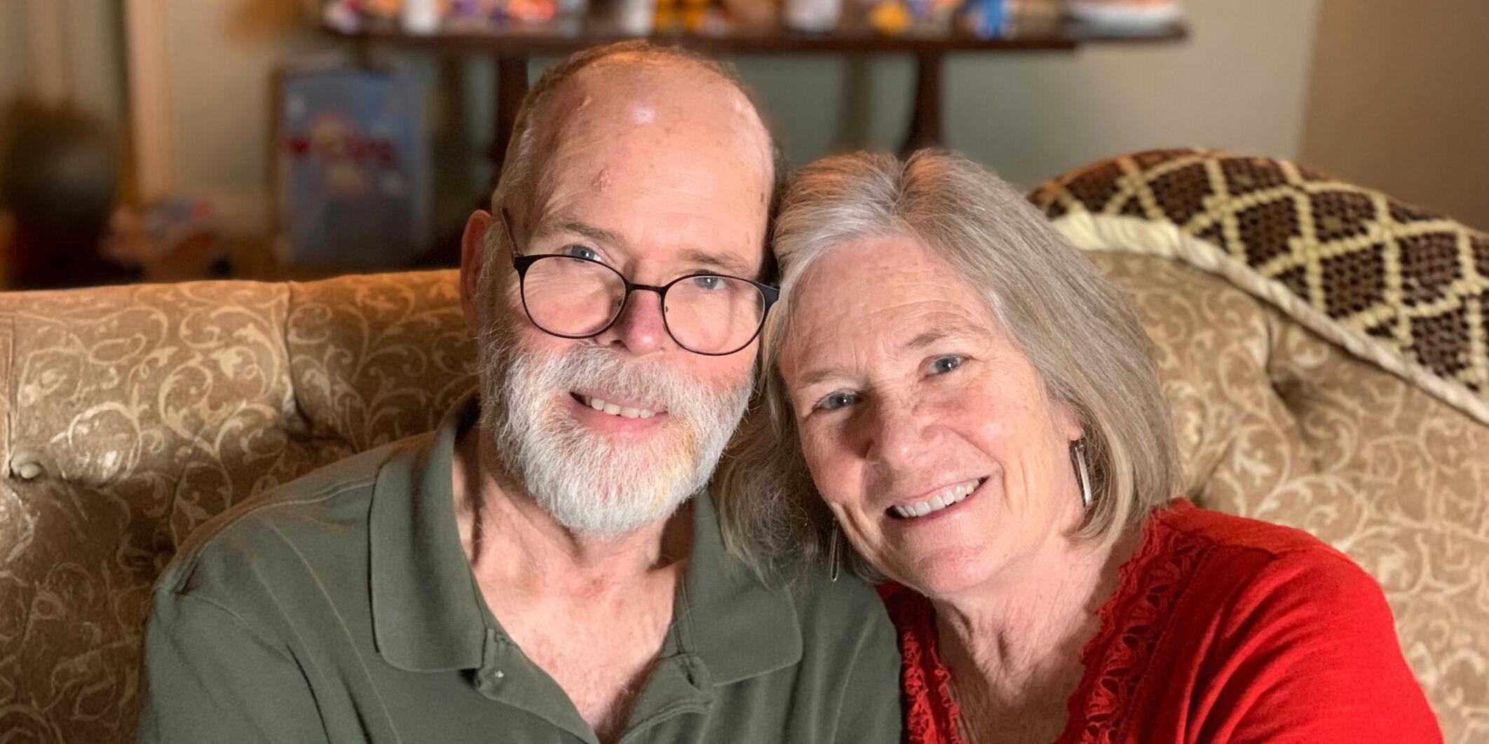 Joanie Prentice and her husband sit and smile together on a couch. 