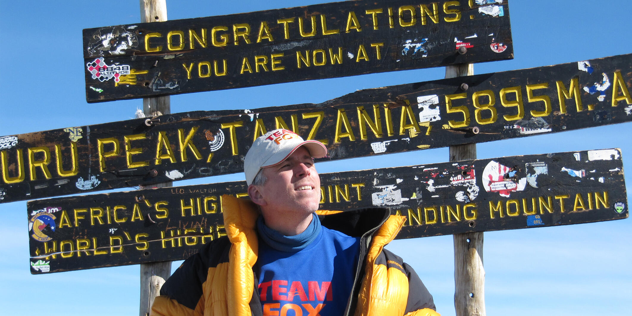 Tom Sabourin stands in front of the sign declaring the Uhuru peak of Mount Kilimanjaro, wearing a Team Fox shirt, and holding a book by Michael J. Fox.